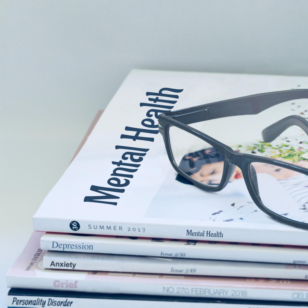 Stack of magazines focused on mental health topics with visible titles such as Mental Health, Depression, Anxiety, Grief, and Personality Disorder from issues dating Summer 2017 and February 2018, accompanied by a pair of black eyeglasses on top.